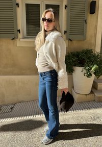 Casaco de pele sintética branca com mangas texturizadas, top bege, jeans boca de sino azuis e ténis cinzentos; segurando uma bolsa preta. Ambiente ao ar livre iluminado pelo sol.