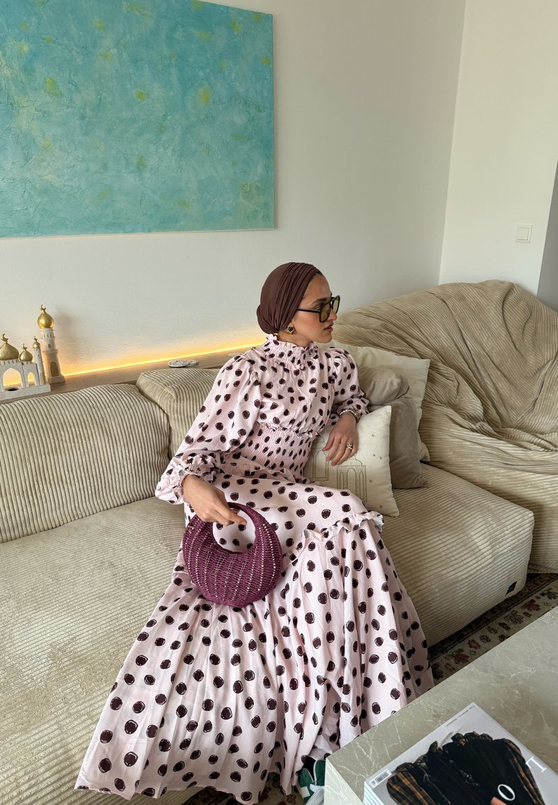 Femme portant un foulard marron et une robe rose à pois, assise sur un canapé beige tenant un sac à main tissé violet, avec une œuvre d'art bleu-vert accrochée au mur.