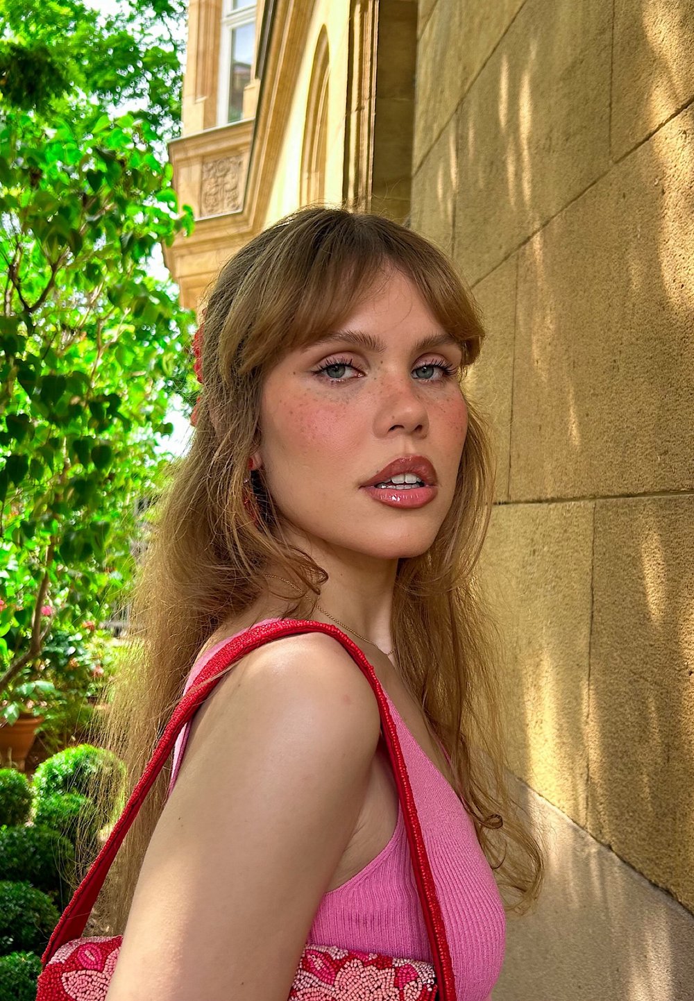 Kvinne iført en rosa ribbet tank top og holder en rød perleveske med blomsterdesign, stående mot en steinmur og grøntområde.