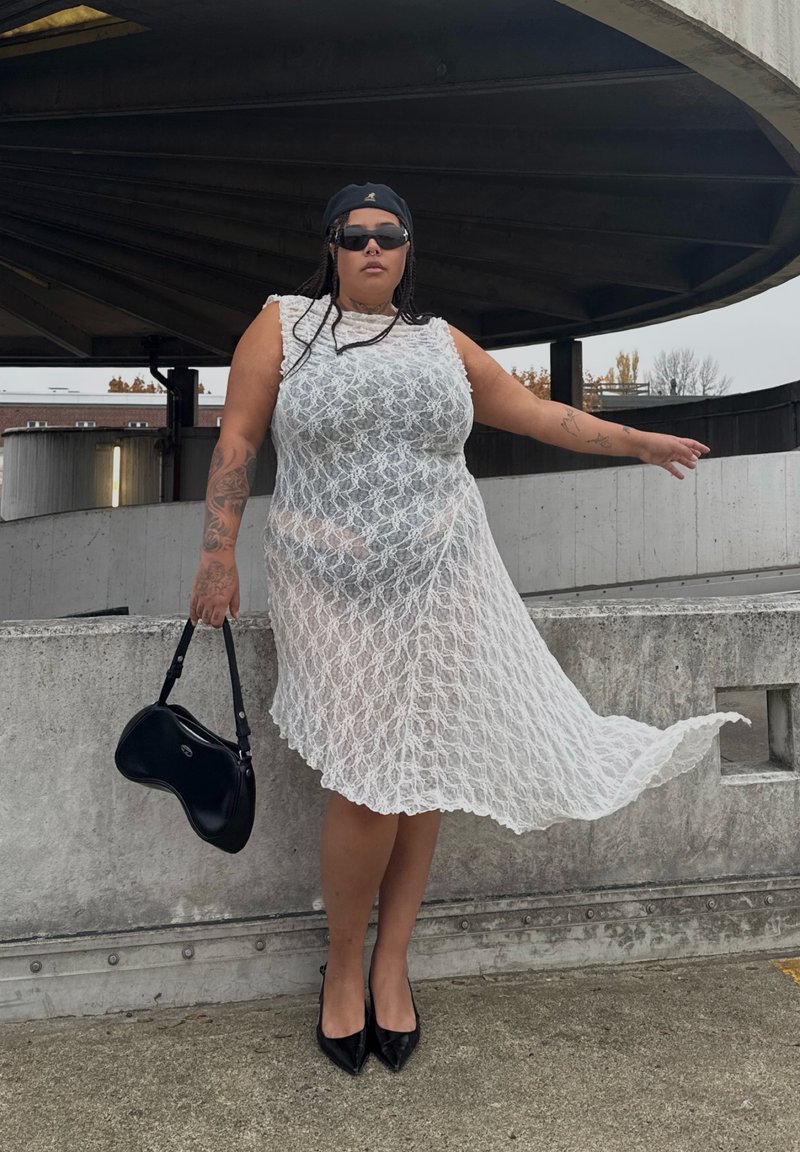 Vestido branco de renda com padrão em ziguezague, design sem mangas e bainha evasê. Mala preta e sapatos de salto alto. Fundo de betão.