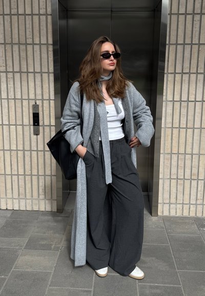 Mujer con el pelo largo, lleva gafas de sol negras, top blanco, cárdigan gris, pantalones anchos oscuros y zapatos blancos, está de pie fuera del ascensor con las manos en los bolsillos.