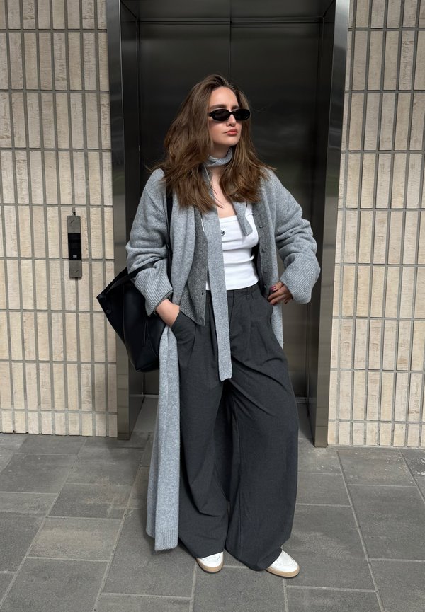 Kvinde med langt hår iført sorte solbriller, hvid top, grå cardigan, vide mørke bukser og hvide sko står udenfor elevatoren med hænderne i lommerne.