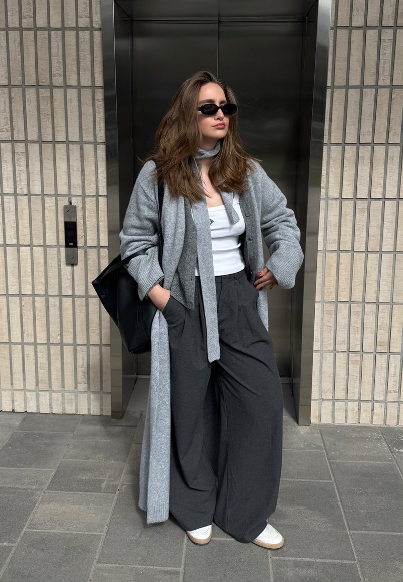 Donna con capelli lunghi che indossa occhiali da sole neri, top bianco, cardigan grigio, pantaloni larghi scuri e scarpe bianche, in piedi fuori dall'ascensore con le mani in tasca.