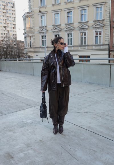 Mujer con chaqueta de cuero marrón oscuro, pantalones holgados y botas, sosteniendo un bolso negro y ajustándose las gafas de sol en una acera urbana.