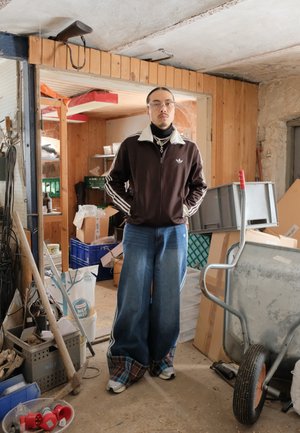 Persona joven con una chaqueta deportiva oscura y jeans anchos, situada en un taller desordenado con una carretilla, cajas y herramientas alrededor.