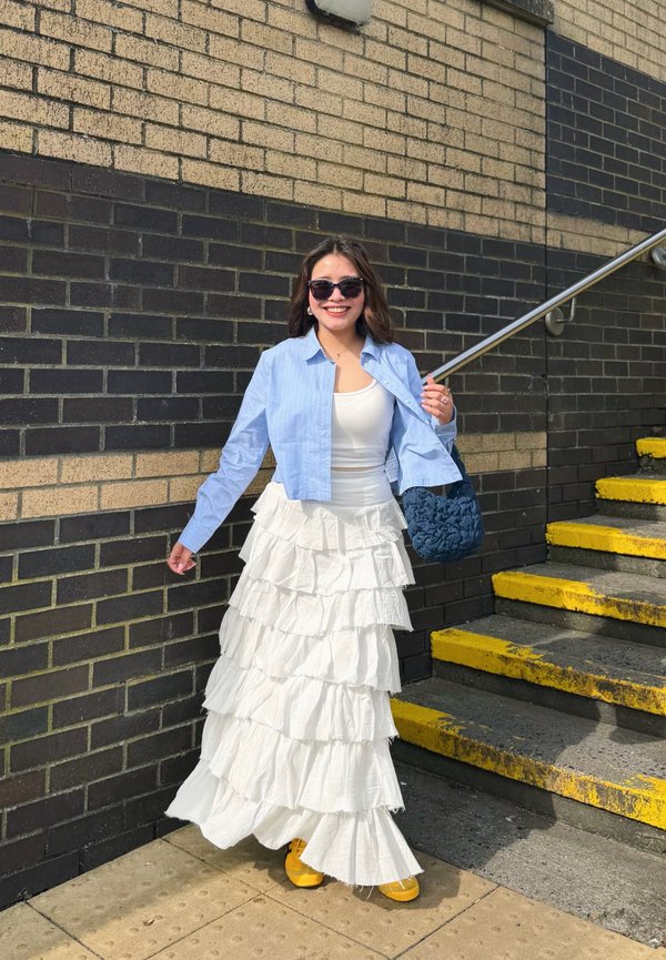 Mujer sonriente con gafas de sol junto a una pared de ladrillos negros y beige al lado de unas escaleras, lleva una camisa azul, falda blanca con volantes y zapatos amarillos.