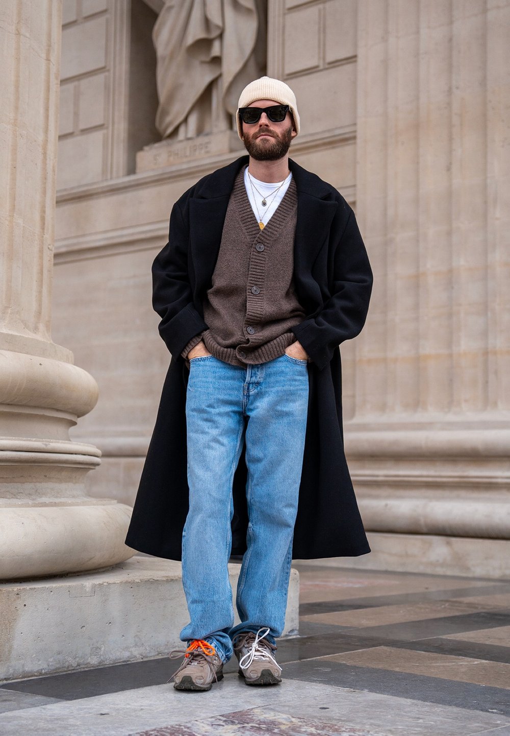 Manteau noir par-dessus un cardigan marron, jean bleu et baskets gris-brun. Bonnet blanc et lunettes de soleil complètent la tenue. Bijoux simples en accents.