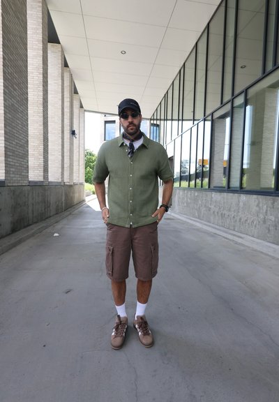Camisa de manga corta verde con textura, pantalones cortos de cargo marrones, calcetines claros, zapatillas de deporte marrones, gorra negra, gafas de sol, de pie en un pasillo.