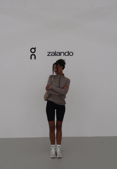 Chaqueta marrón claro con cremallera sobre pantalones cortos negros de ciclismo, combinada con zapatos deportivos blancos y calcetines hasta el tobillo, de pie contra una pared blanca lisa.