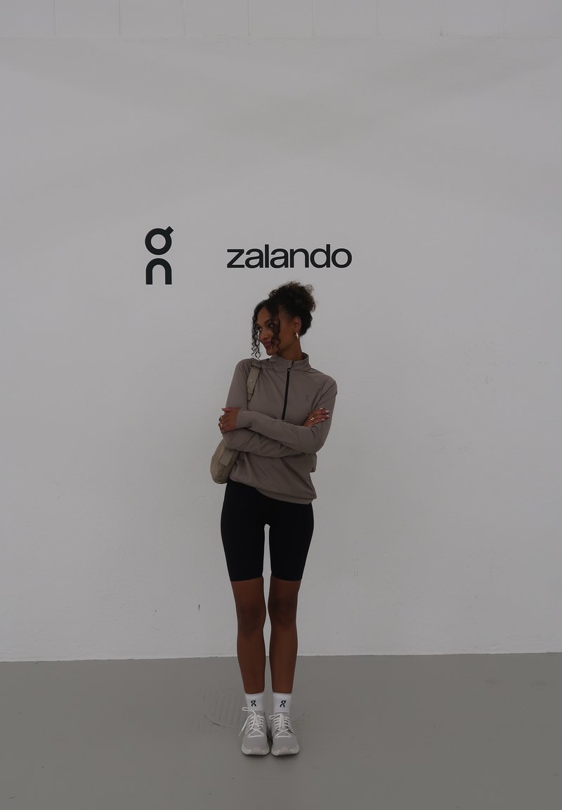 Light brown zip-up jacket over black cycling shorts, paired with white athletic shoes and ankle socks, standing against a plain white wall.