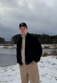 Jeune homme portant une casquette noire, une veste noire, un pull beige et un pantalon, debout près d'une rive enneigée avec une forêt et un ciel nuageux en arrière-plan.