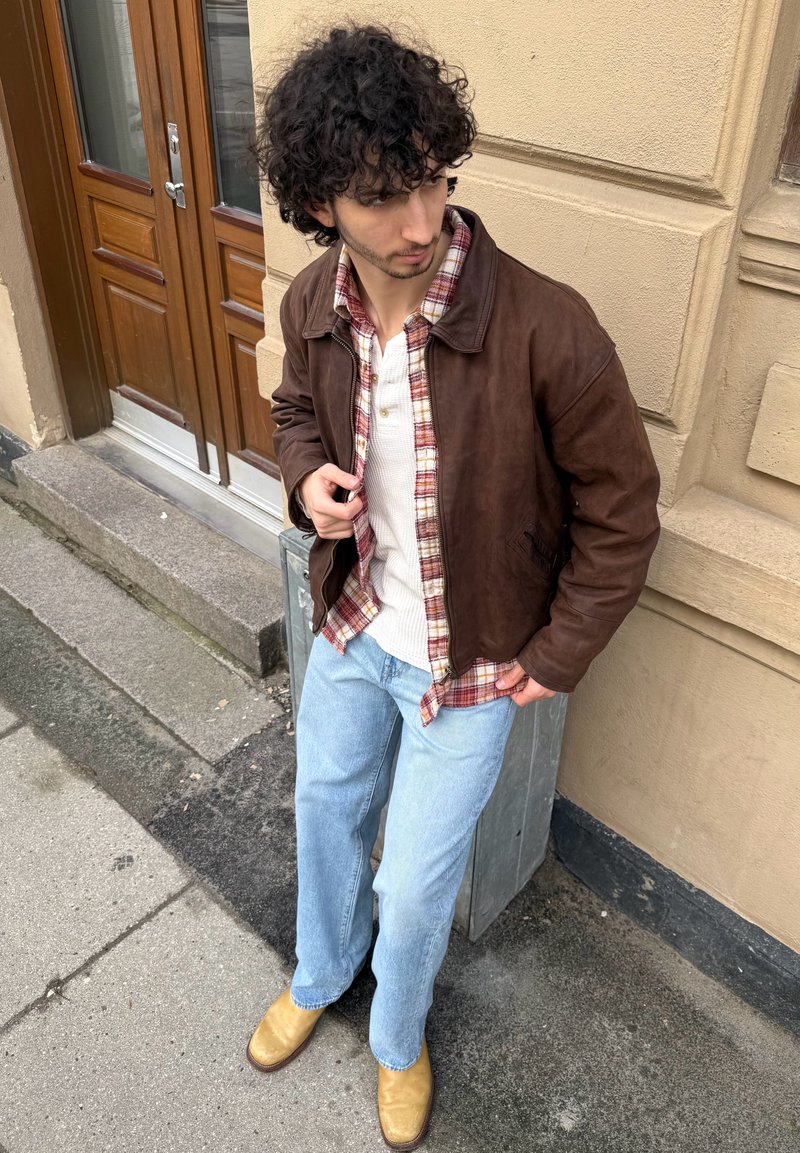 Jeune homme aux cheveux bouclés portant une veste en cuir marron, une chemise à carreaux, un henley blanc, un jean bleu clair et des bottes couleur beige, debout près d'un mur beige à côté d'une porte en bois.