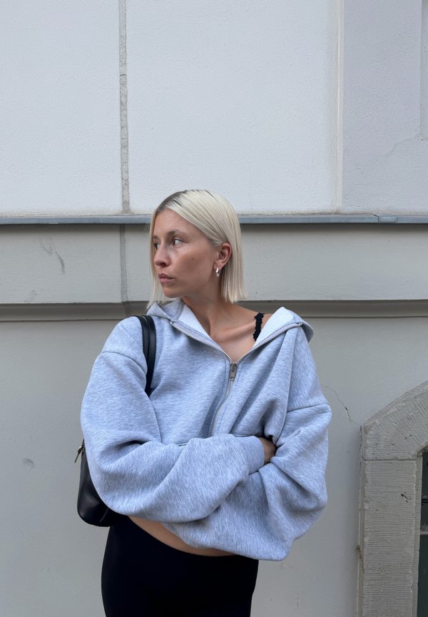 Kvinna med kort blont hår som bär en grå oversized zip-hoodie och svart topp, stående med armarna i kors nära en beige vägg.