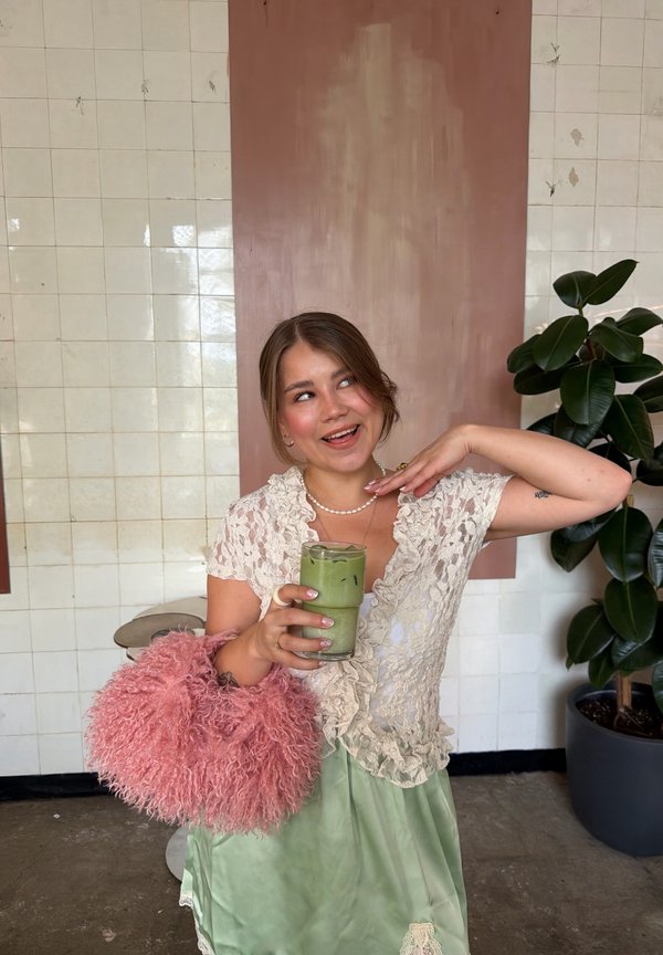Jonge vrouw in kanten top en groene rok houdt ijskoude matcha drank en roze pluizige tas vast, staand bij plant en betegelde muur.