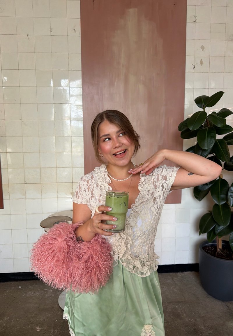 Jeune femme en haut en dentelle et jupe verte tenant une boisson matcha glacée et un sac rose duveteux, debout près d’une plante et d’un mur carrelé.