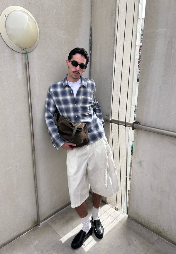 Hombre apoyado en la esquina de una pared, con gafas de sol, camisa de cuadros, pantalones cortos blancos, zapatos negros, sosteniendo una bolsa marrón en el balcón.