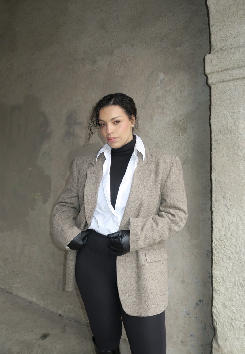 Kvinne med krøllete hår iført hvit skjorte, svart høyhalset genser, beige blazer, svarte hansker og svarte bukser, stående mot en strukturert vegg.