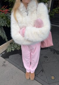 Bílá bunda z umělého kožíšku s růžovými rukávy, světle růžové kalhoty a béžové kotníkové boty. Doplňováno růžovou kabelkou a postava stojí na dlaždicové ploše.