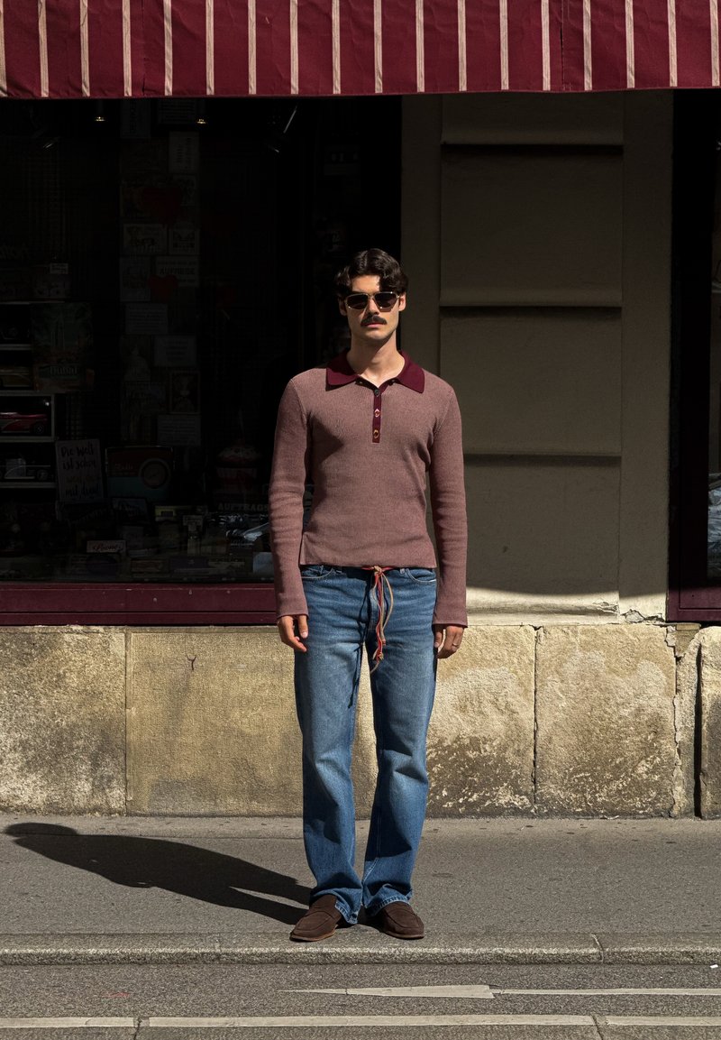 Sweater de gola com mangas compridas em bordeaux e cinza, com um acabamento texturizado, combinado com jeans azuis e sapatos castanhos, usados ao ar livre.