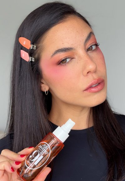 Mujer con sombra de ojos rosa y clips en el cabello sostiene un frasco de spray Sol de Janeiro 16-Hour Setting Mist sobre un fondo gris liso.