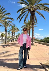 Gestreepte roze oversized shirt, witte t-shirt, donkere wijde jeans en zwarte sandalen. Palmbomen en blauwe lucht op de achtergrond.