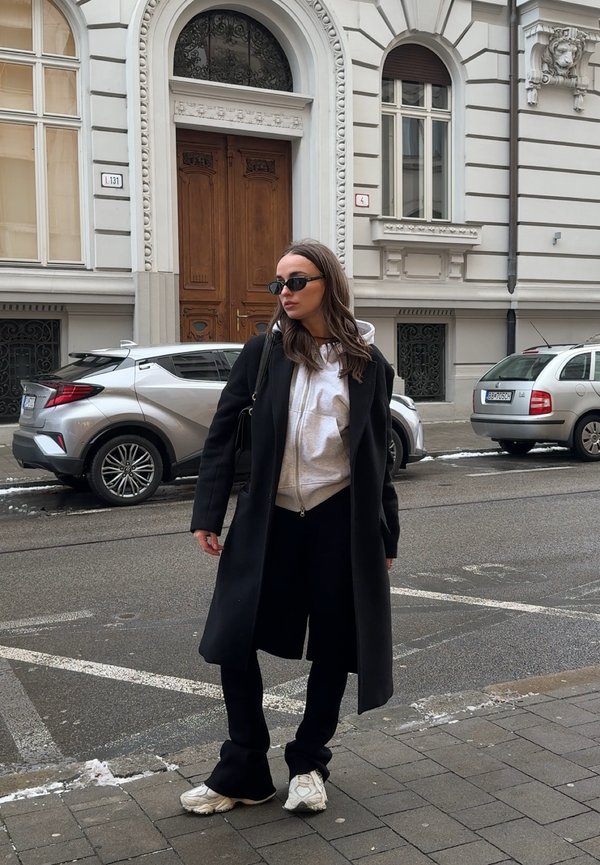 Vrouw met zwarte jas, witte hoodie, zwarte broek en witte sneakers staat op een stadsstoep met auto's en een sierlijk gebouw op de achtergrond.