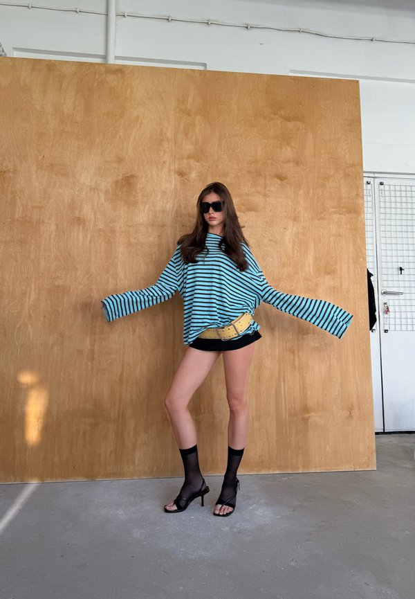 Donna che indossa una camicia oversize a righe blu e nere, pantaloncini neri, calzini neri trasparenti, sandali con tacco alto, occhiali da sole, in piedi davanti a un pannello di legno.