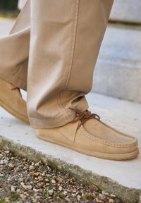 Beige ruskinds mokkasin-sko med rund tå, snøredetalje og gummisål, båret med brune bukser på en grusoverflade.