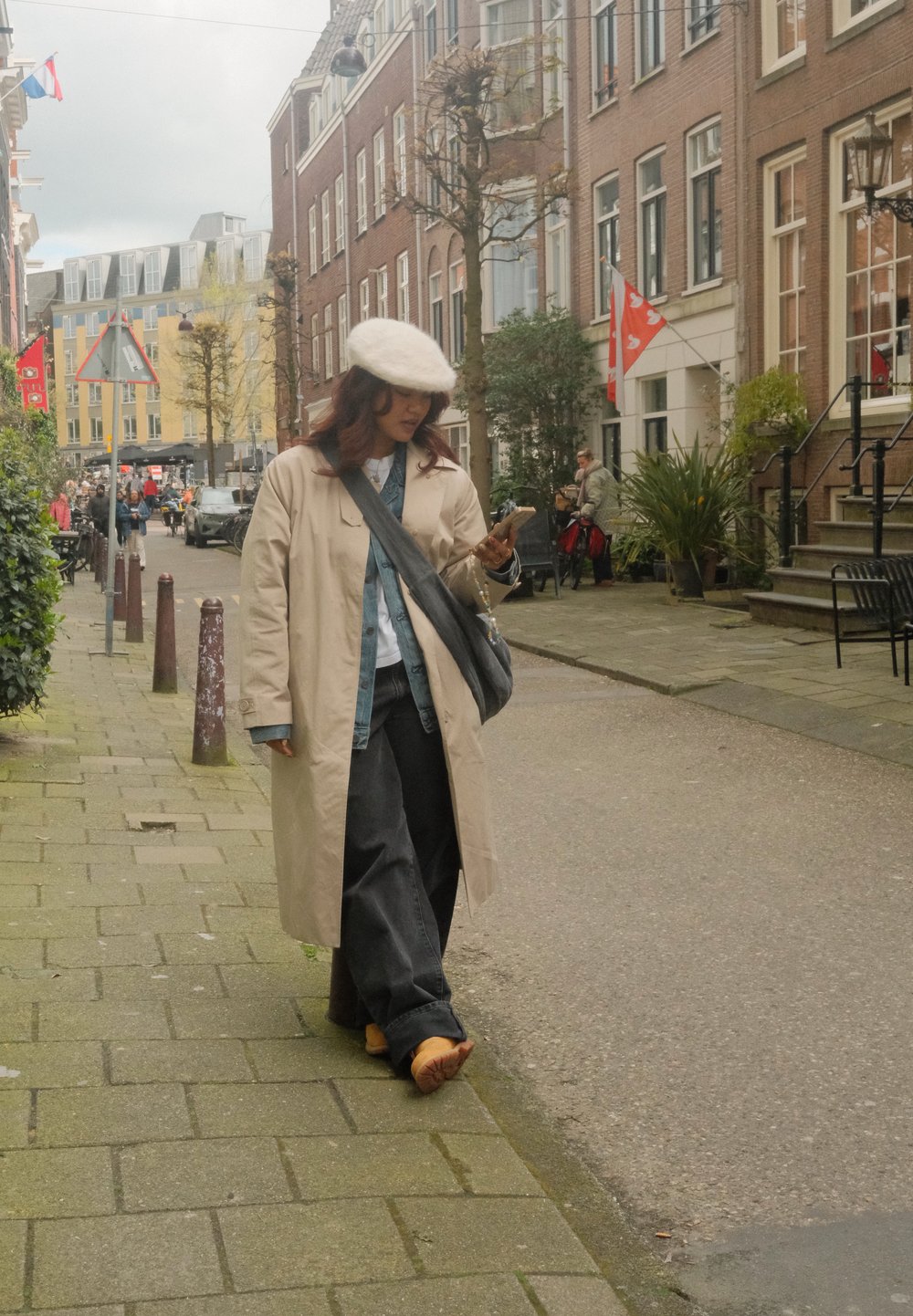 Beige lang jakke over en blå denimskjorte, mørke bukser og brune sko, som går på en brosteinsgate og ser på en smarttelefon.