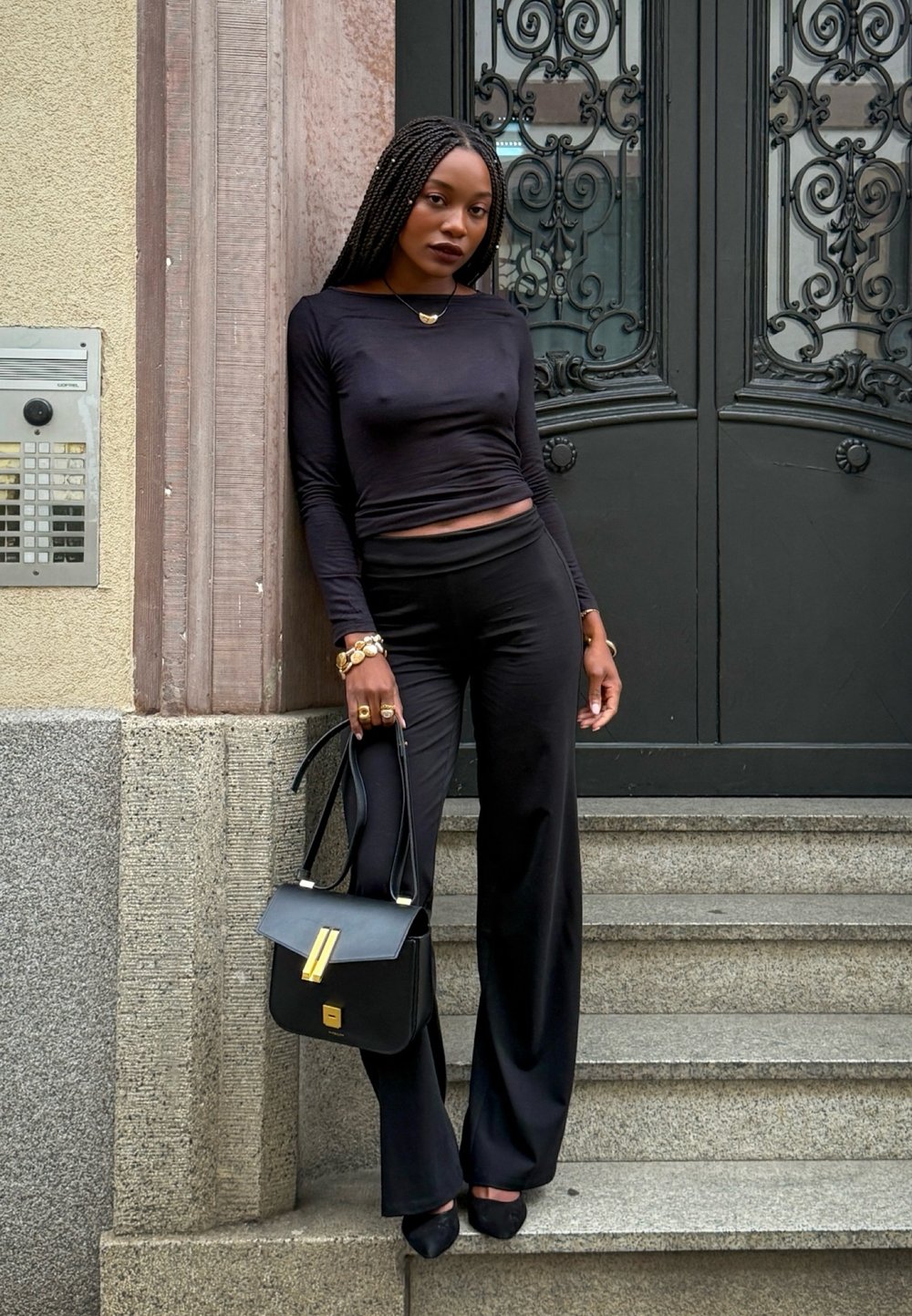 Zwarte cropped top met lange mouwen, zwarte high-waisted broek, zwarte handtas met gouden accenten, gedragen met zwarte hakken, staande bij ornamentele deuren en trappen.