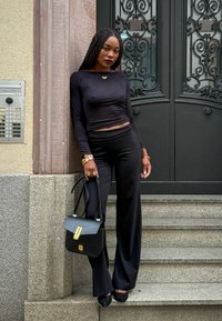 Zwarte cropped top met lange mouwen, zwarte high-waisted broek, zwarte handtas met gouden accenten, gedragen met zwarte hakken, staande bij ornamentele deuren en trappen.