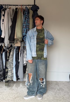 Hombre con camisa de camuflaje, chaqueta ligera de mezclilla, jeans anchos con roturas y zapatos tipo slip-on a cuadros, de pie junto a un perchero con chaquetas y camisas.