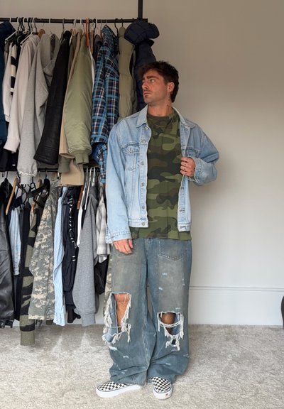 Hombre con camisa de camuflaje, chaqueta ligera de mezclilla, jeans anchos con roturas y zapatos tipo slip-on a cuadros, de pie junto a un perchero con chaquetas y camisas.