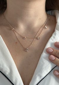 Collana a più strati in tono oro con charm a forma di farfalla e un fiore centrale con gemme. Indossata sopra una camicia bianca con colletto.