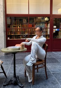 En person i en lys grå treningsdrakt sitter på en trestol ved et rundt bord med metalltoppen, holder en drink og et magasin.