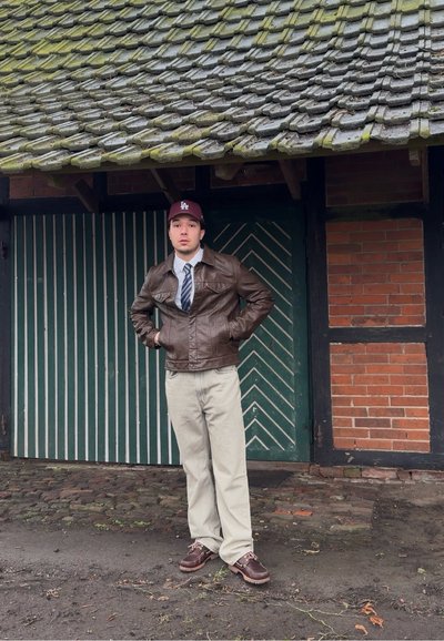 Joven de pie con las manos en los bolsillos, vistiendo una chaqueta de cuero marrón, corbata, pantalones beige y gorra granate bajo un techo de tejas cubierto de musgo.