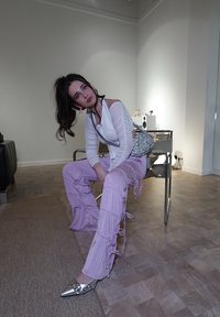 Ανοιχτό μωβ, ριγμένες παντελόνες με κορδόνια και εφαρμοστό λευκό μπλουζάκι με μακριά μανίκια. Ασημένια flats με μυτερή μύτη και τσάντα με ασημένια παγιέτα. Μινιμαλιστική διακόσμηση.