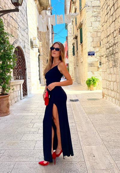 Vestido maxi negro con una abertura alta hasta el muslo, combinado con tacones rojos y un bolso a juego. Calle de piedra y edificios en el fondo.