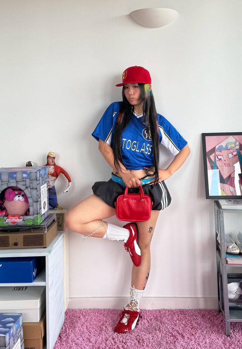 Jeune femme en maillot de sport bleu, jupe noire, casquette rouge et chaussures rouges pose avec un sac à main rouge dans une pièce avec des jouets et des œuvres d'art encadrées.