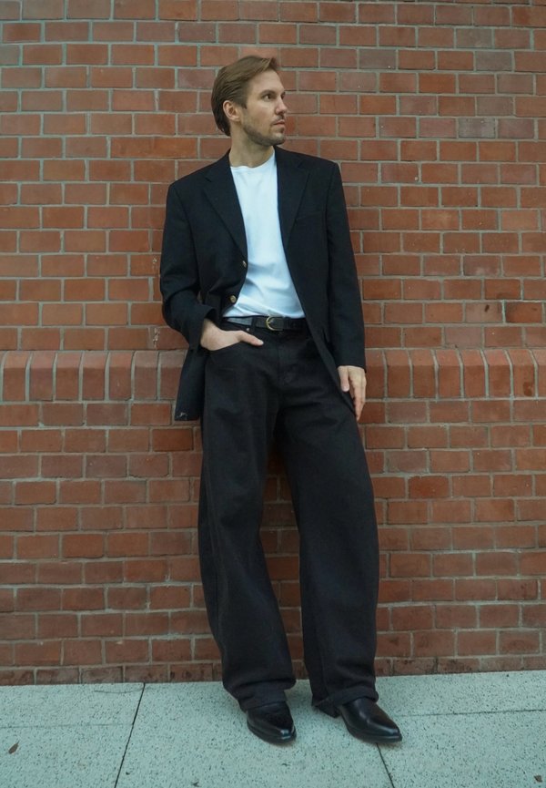 Uomo in giacca nera, pantaloni larghi neri, camicia bianca e scarpe nere, appoggiato a un muro di mattoni rossi con una mano in tasca.