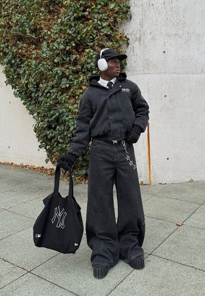 Chaqueta de mezclilla negra oversize con capucha, pantalones anchos negros, guantes negros y una bolsa tote negra con un logo blanco de NY. Auriculares blancos.