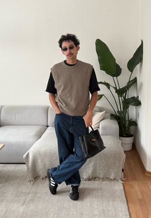 Hombre con gafas de sol, camiseta negra, suéter sin mangas marrón, vaqueros y zapatillas negras, de pie en el interior sosteniendo un bolso negro cerca de un sofá y una planta.