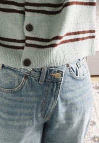 Jeans a vita alta di colore azzurro chiaro con una texture sbiadita, abbinati a un cardigan verde menta con strisce marrone scuro e chiusure con bottoni.
