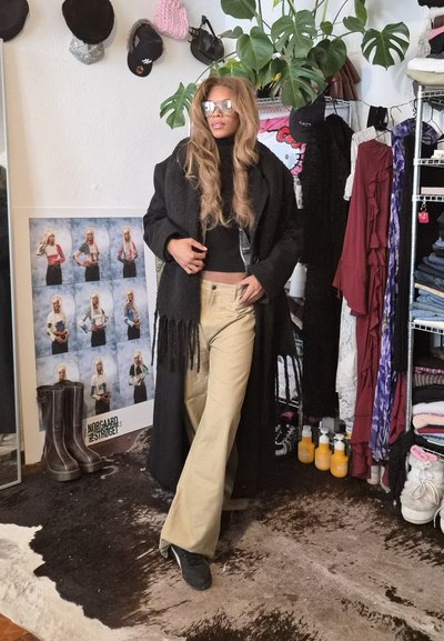Mujer con cabello largo, vistiendo un abrigo negro, pantalones beige y gafas de sol, de pie en una habitación desordenada con ropa, plantas y botas.