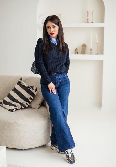 Mujer con cabello largo y oscuro, vestida con un suéter azul marino, jeans azules y zapatillas deportivas, de pie junto a un sofá beige con cojines a rayas en una habitación luminosa.