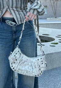 Λευκή υφασμάτινη τσάντα ώμου με ασημί εξαρτήματα, διαθέτει φερμουάρ και διακοσμητικά τ studs, κρατιέται από ένα άτομο που φορά τζιν και καρό πουκάμισο.