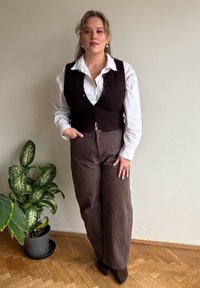 Brown waistcoat over a white button-up shirt, paired with wide brown trousers and pointed brown boots. Indoor setting with a green plant.