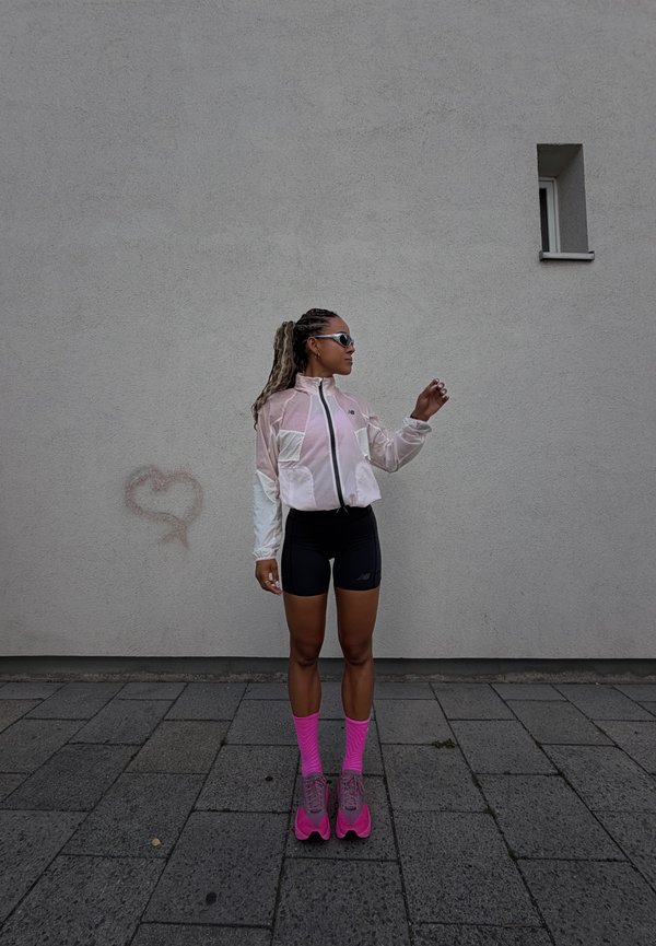 Persona con giacca rosa e bianca, pantaloncini neri, calzini rosa acceso e scarpe da ginnastica in piedi sul marciapiede contro un muro grigio con una piccola finestra.