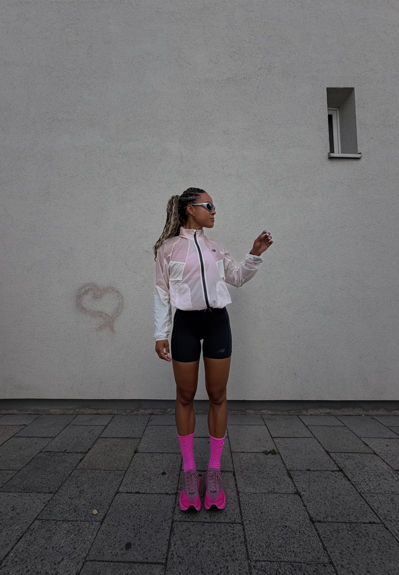 Personne en veste rose et blanche, short noir, chaussettes rose vif et baskets, debout sur le trottoir contre un mur gris avec une petite fenêtre.