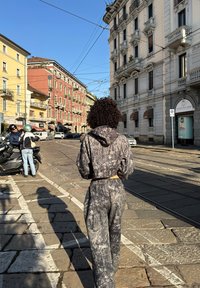 Kiharatukkainen nainen harmaassa kuvioidussa verkkaritakissaan kävelee aurinkoisella kaupunkikadulla, jonka varrella on potkulautoja ja värikkäitä rakennuksia.